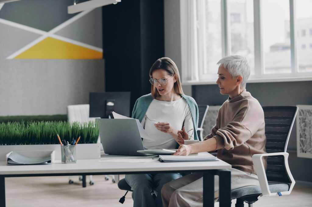 Two confident women using technologies and communicating while working in office together